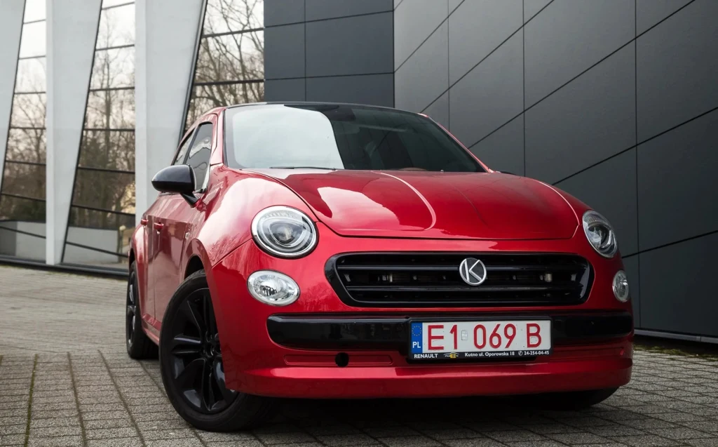 Czerwony samochód osobowy o nowoczesnym wyglądzie stoi zaparkowany na tle szklanej i metalowej elewacji budynku, z tablicą rejestracyjną i czarnymi felgami, światła przednie oraz linie karoserii dobrze widoczne.