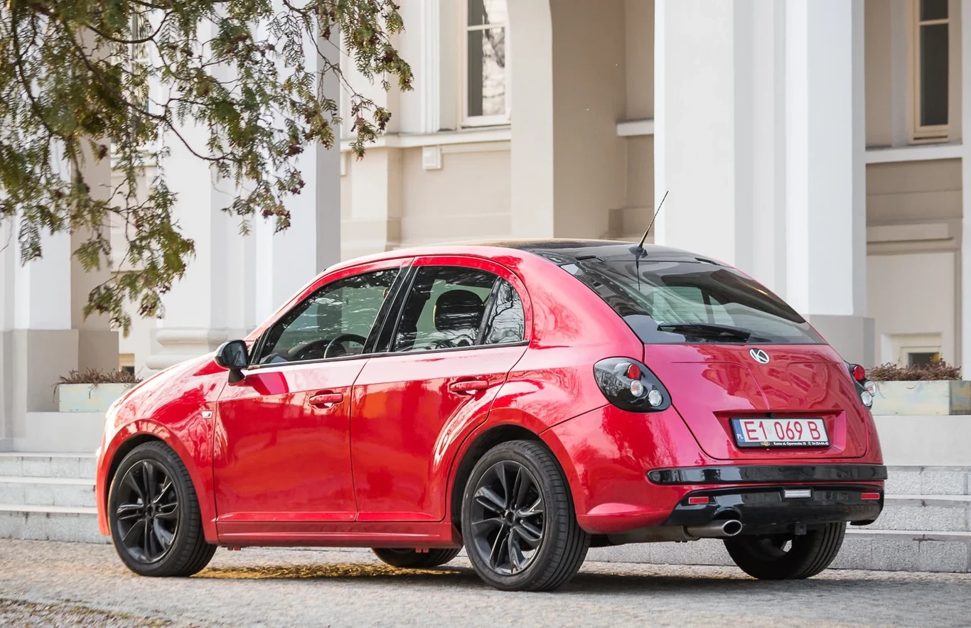 Czerwony samochód osobowy zaparkowany na brukowanym podjeździe przed budynkiem z białymi kolumnami, widoczne czarne felgi, tablica rejestracyjna oraz fragment drzewa po lewej stronie kadru.