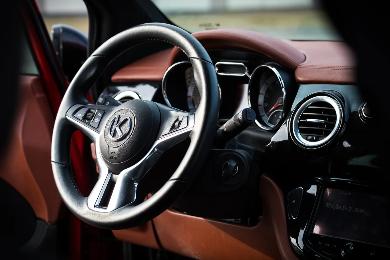 Wnętrze samochodu z widoczną kierownicą, zegarami i deską rozdzielczą wykończoną brązową skórą, elementy czarne i chromowane, dobrze doświetlone światłem dziennym.