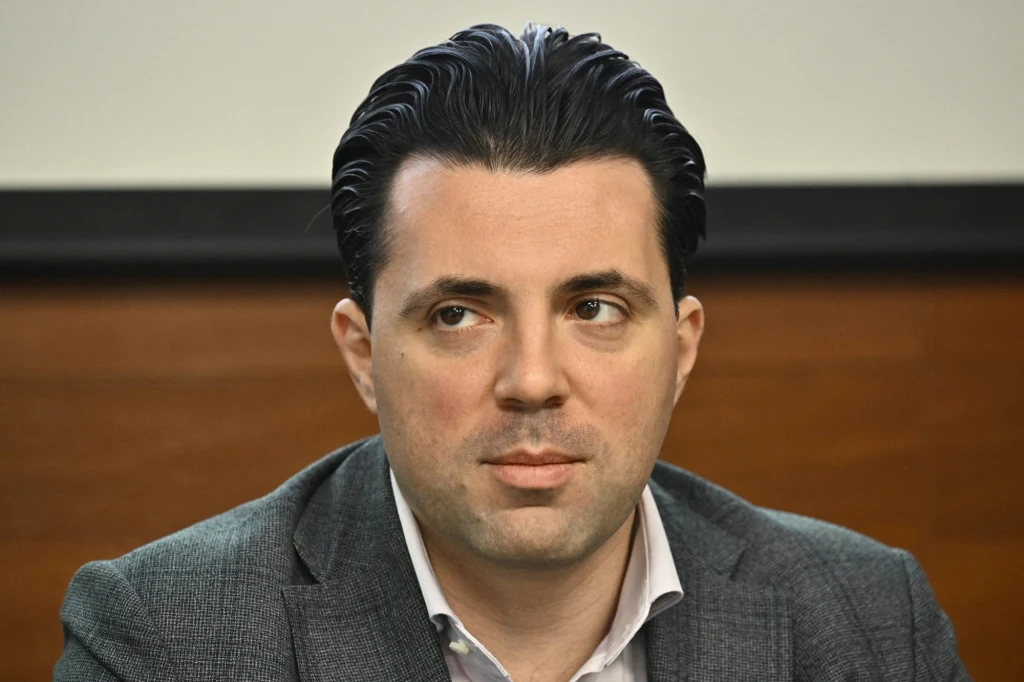 Mężczyzna w szarej marynarce i białej koszuli siedzi na tle brązowej ściany, z zaczesanymi do tyłu włosami, patrzy lekko w bok, wyrażając skupienie i powagę.