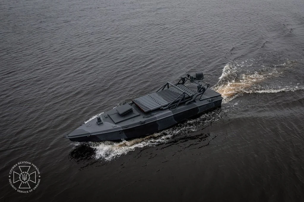 Czarna, nowoczesna łódź wojskowa płynie po spokojnej powierzchni wody, wzburzając fale. W lewym dolnym rogu widoczne jest logo instytucji.