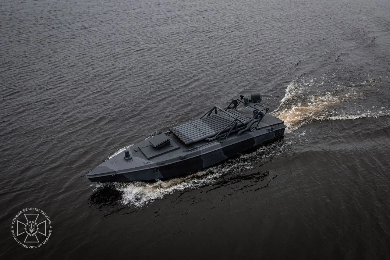 Czarna, nowoczesna łódź wojskowa płynie po spokojnej powierzchni wody, wzburzając fale. W lewym dolnym rogu widoczne jest logo instytucji.