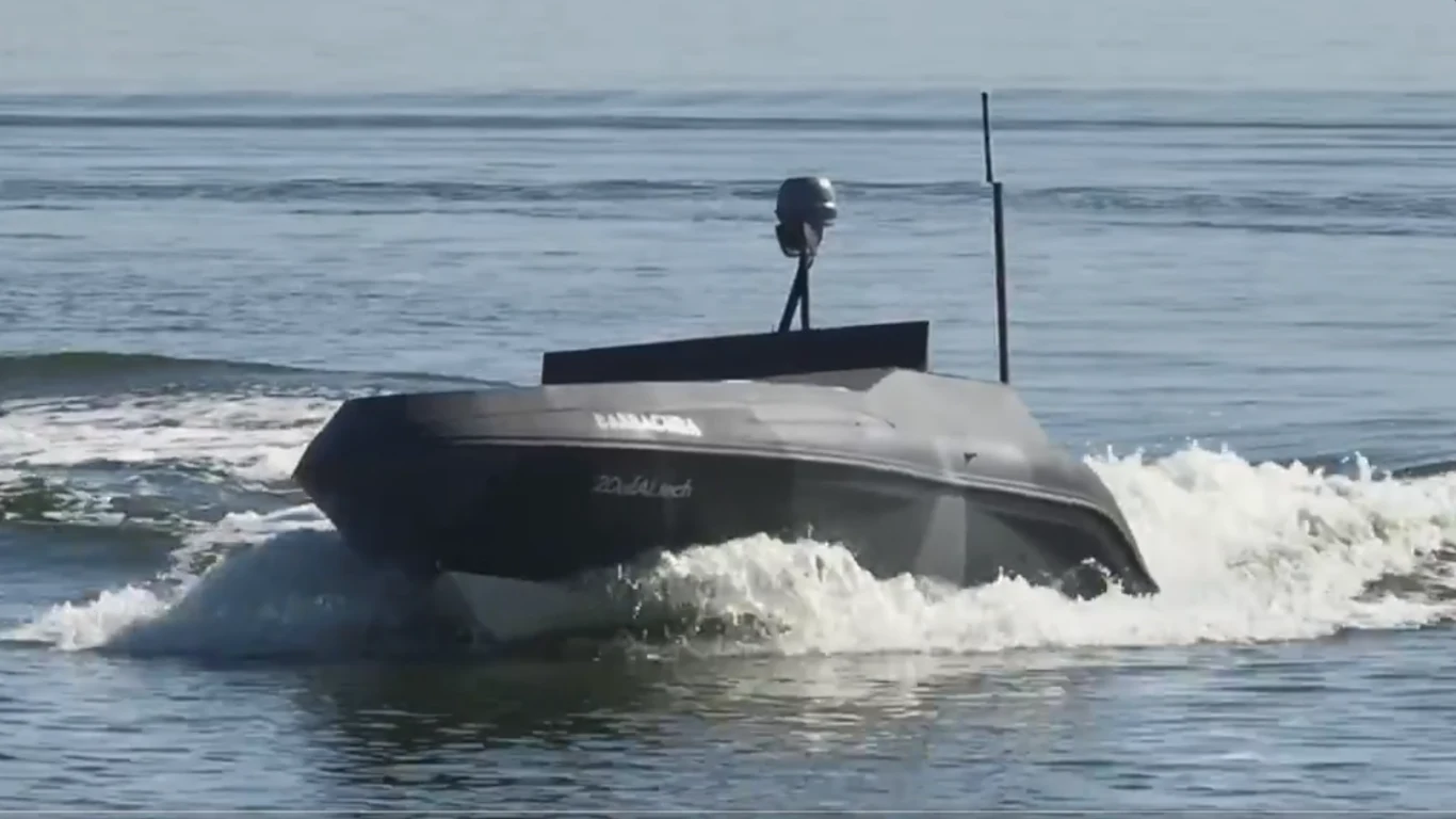 Bezzałogowa łódź poruszająca się po spokojnej powierzchni wody, wywołująca fale na swoim torze. Kadłub pojazdu jest opływowy, ciemny, z widoczną anteną i kamerą na szczycie.