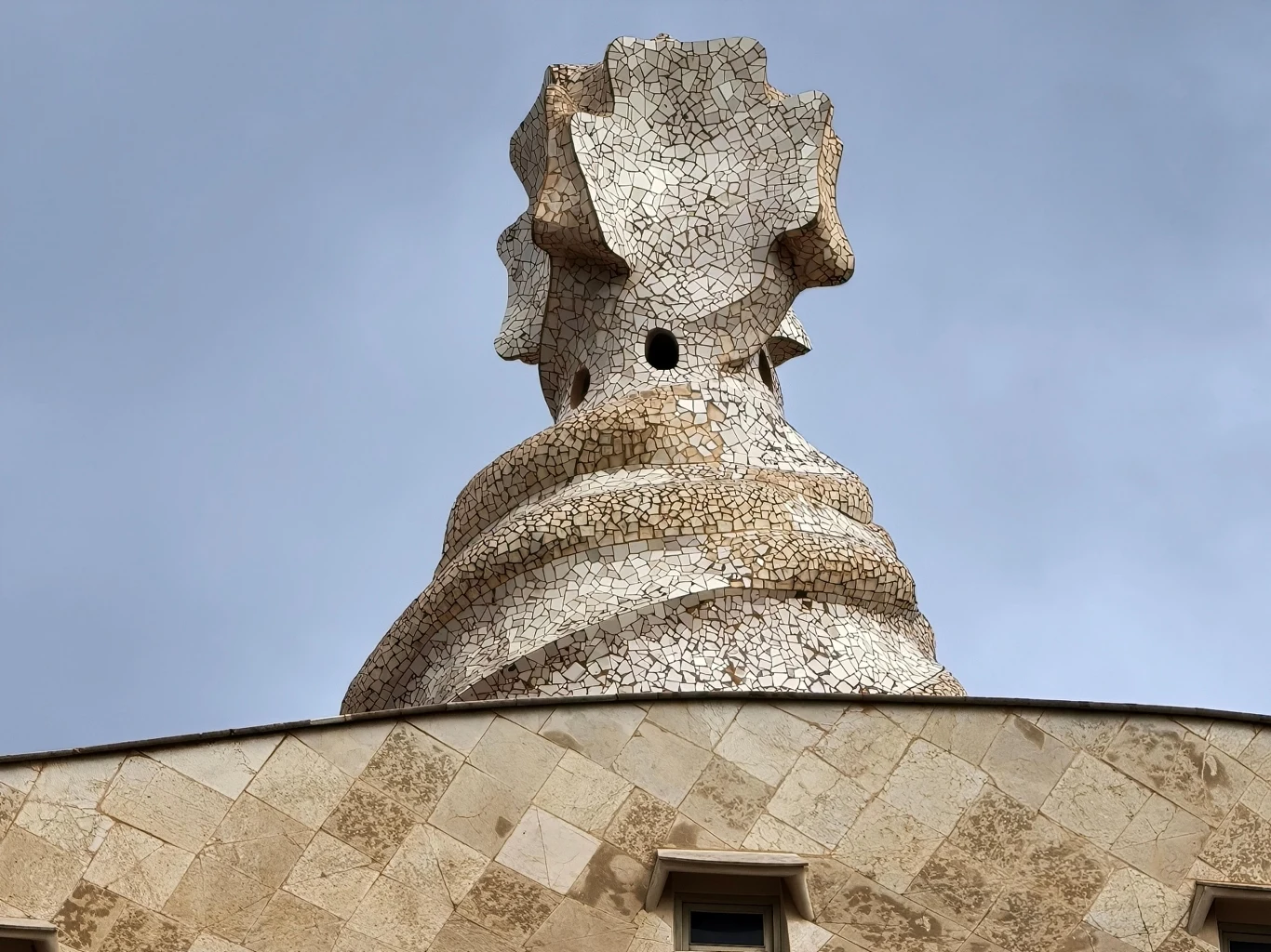 Fragment nieregularnej, mozaikowej kompozycji na dachu budynku, zdominowany przez oryginalną, spiralną rzeźbę przypominającą fantazyjny komin lub wierzę obserwacyjną. Struktura pokryta jest spękaną ceramiką w jasnej kolorystyce. Tło wypełnia szare niebo.