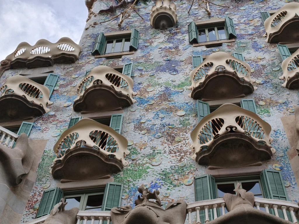 Casa Batlló w Barcelonie. Fasada ozdobnego budynku z unikalnymi balkonami o organicznych kształtach, kolorowymi mozaikami i zielonymi okiennicami, wyróżniająca się artystycznym stylem architektonicznym.