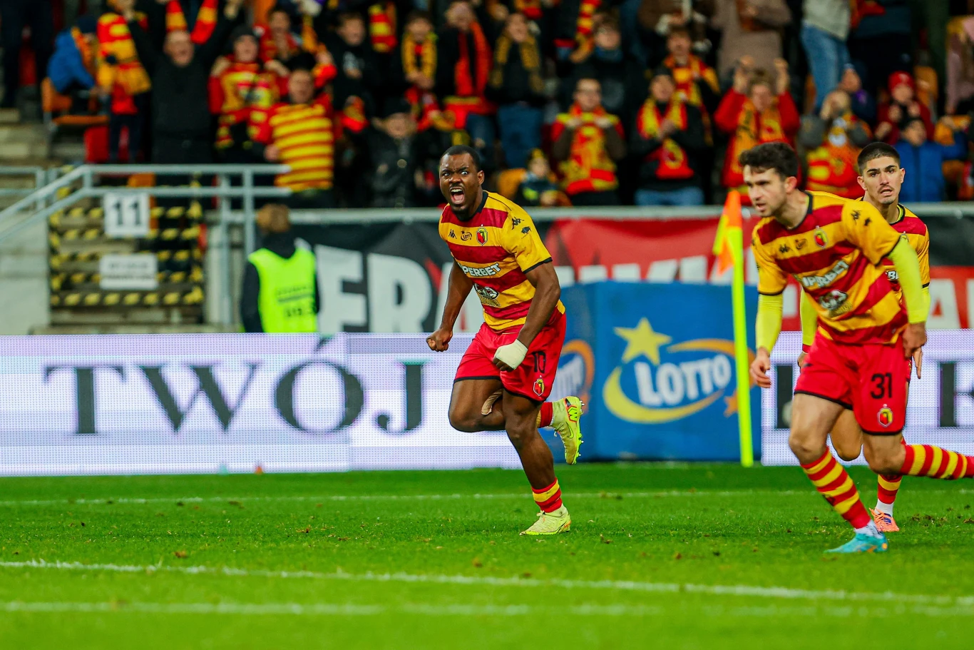 Afimico Pululu i Leon Flach w barwach Jagiellonii Białystok Piłkarze w żółto-czerwonych strojach świętują zdobycie gola na stadionie, kibice w tle również ubrani w klubowe barwy cieszą się i dopingują swoją drużynę.