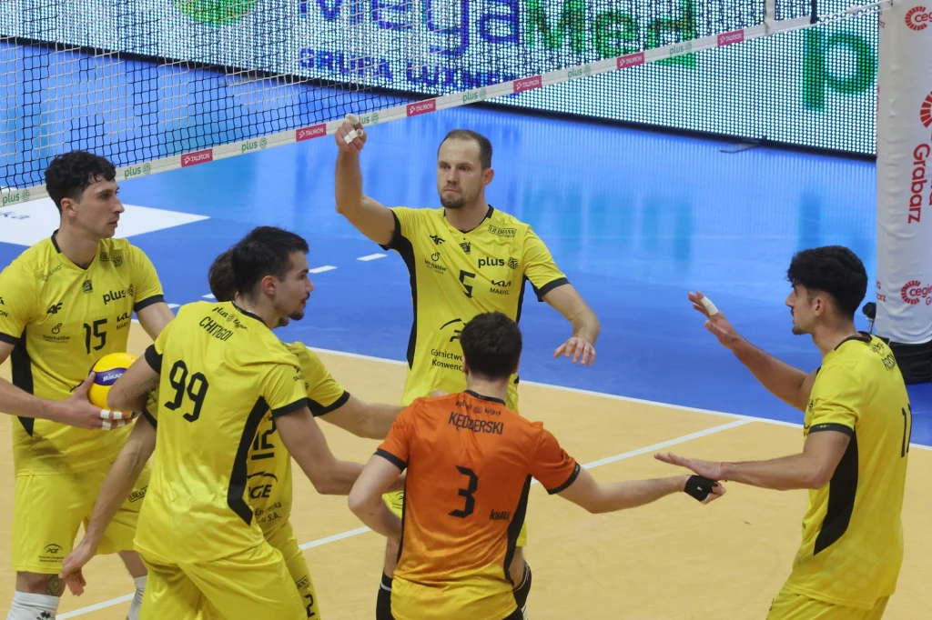 Siatkarze PGE GiEK Skry Bełchatów Grupa siatkarzy ubranych w żółte stroje drużyny zgromadzona wokół zawodnika w pomarańczowej koszulce, unoszą ręce i wymieniają gesty podczas meczu na hali sportowej, w tle widoczna siatka oraz reklamy.