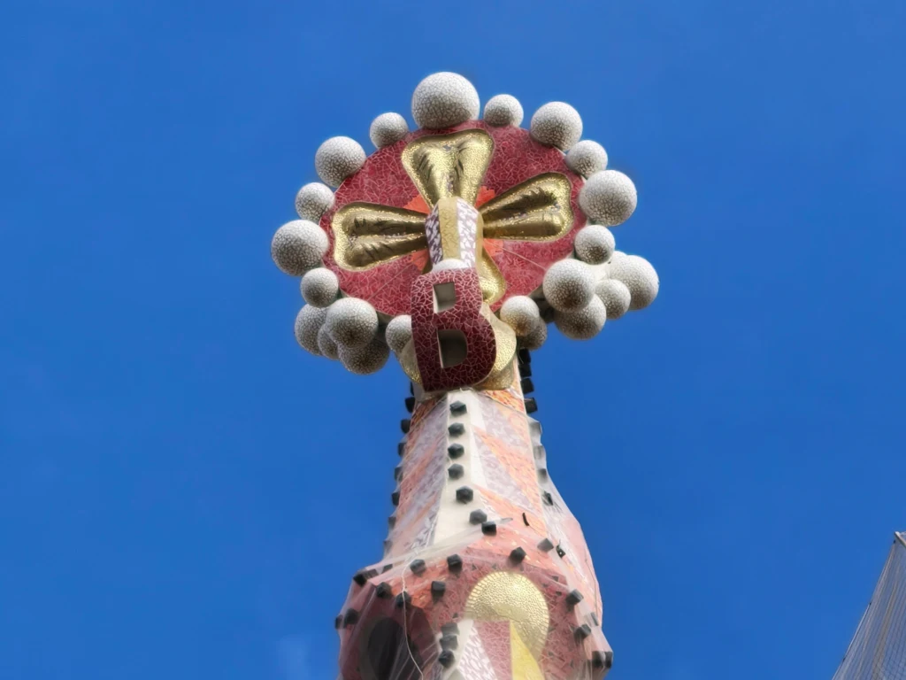 Krzyż na wieży Sagrady Familii w Barcelonie na 20-krotnym zblizeniu. Szczyt wieży ozdobiony kolorową mozaiką z widocznym dużym krzyżem otoczonym okrągłymi elementami oraz czerwoną literą B, na tle błękitnego nieba.