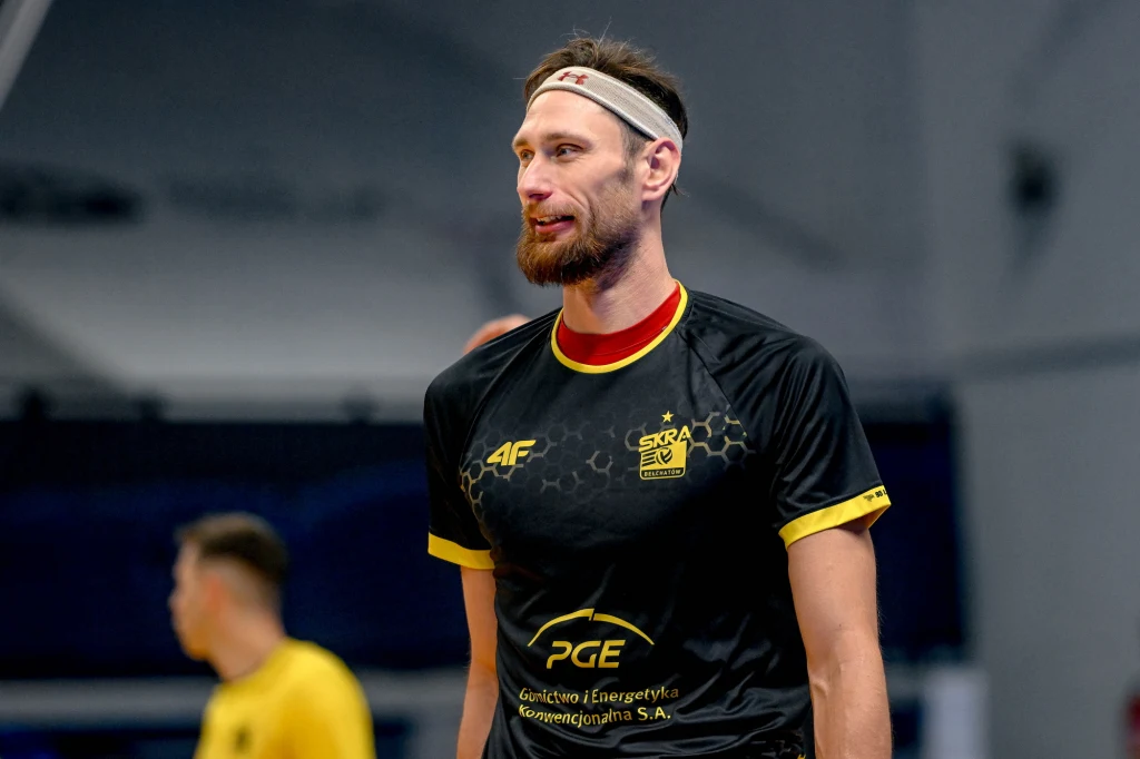 Siatkarz w czarnej koszulce sportowej z żółtymi akcentami i logotypami sponsorów, z opaską na głowie, stoi na hali sportowej, w tle widoczna druga postać w żółtym stroju.