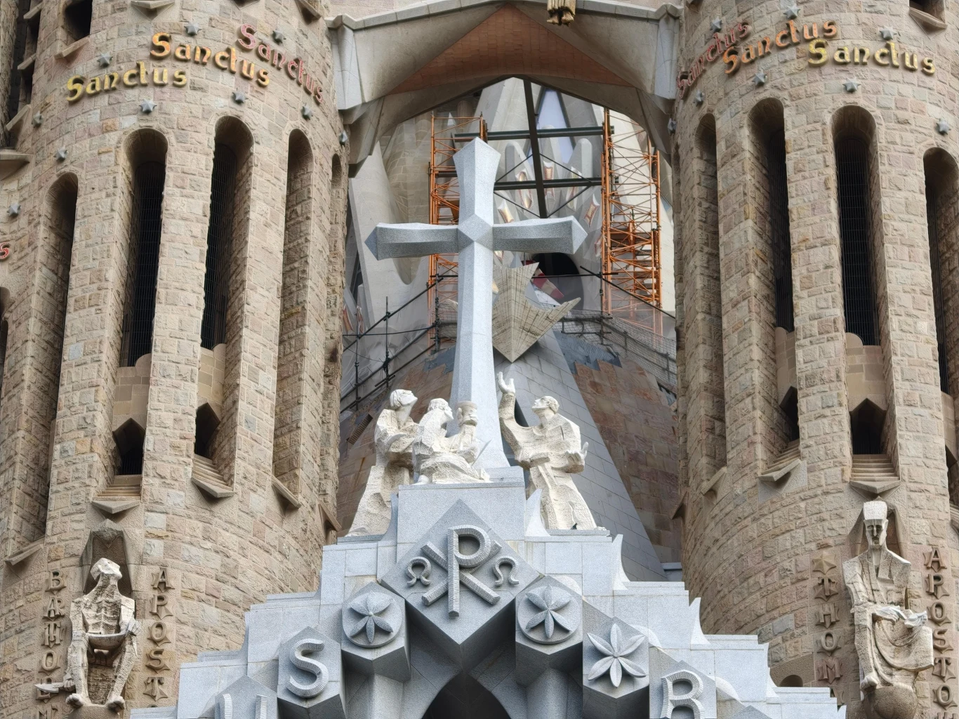 Kamienna fasada z trzema wysokimi wieżami, zdobiona rzeźbami, dużym krzyżem oraz napisem 'Sanctus' na wieżach, charakterystyczna dla Sagrada Família w Barcelonie.