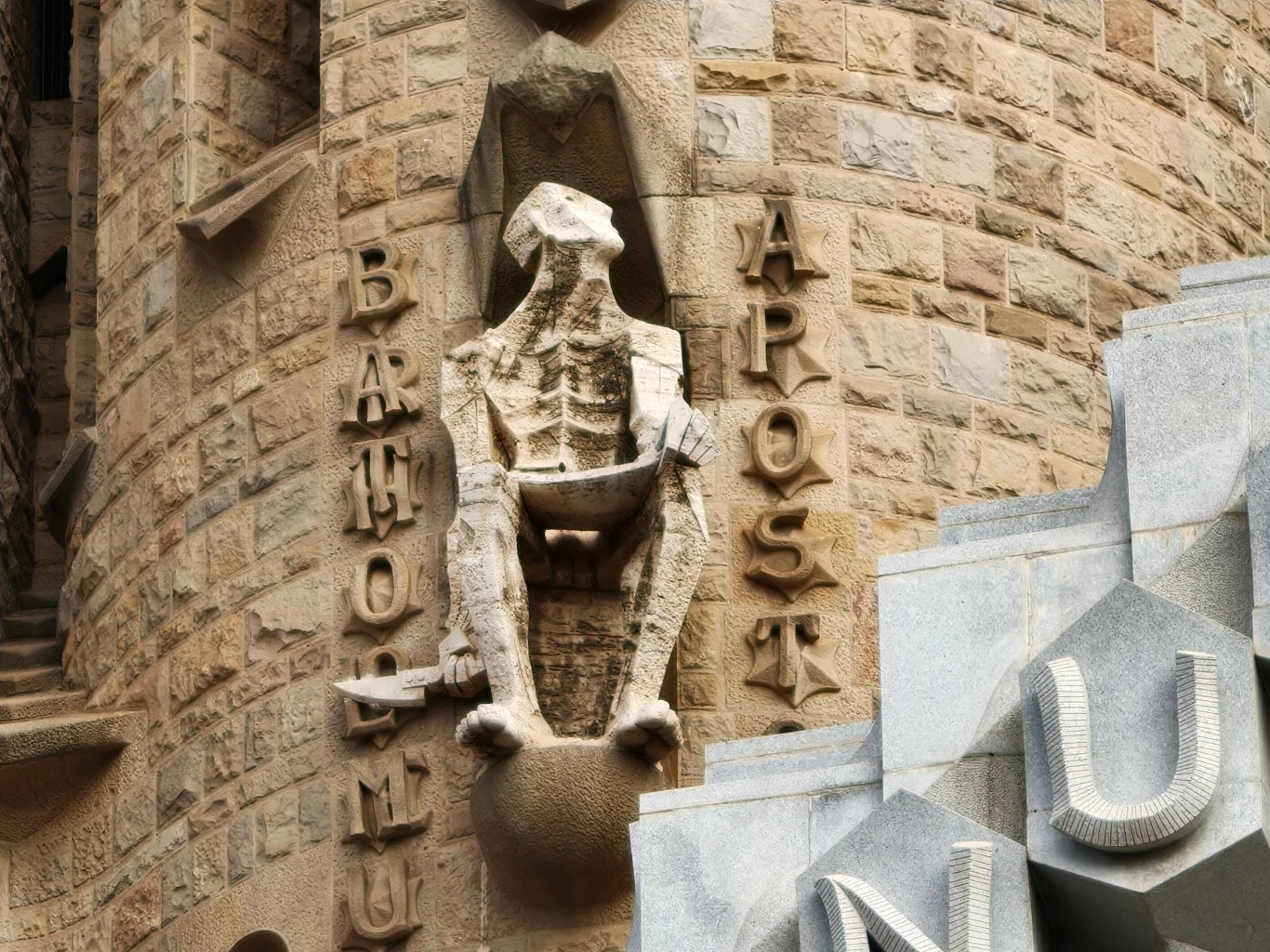 Kamienna rzeźba apostoła Bartłomieja o szkieletowej sylwetce, umieszczona na ścianie budowli, otoczona napisem z wyrazami 'BARTHOLOMEU' i 'APOSTOL' w pionowym układzie liter, wyrazista tekstura kamiennej ściany.