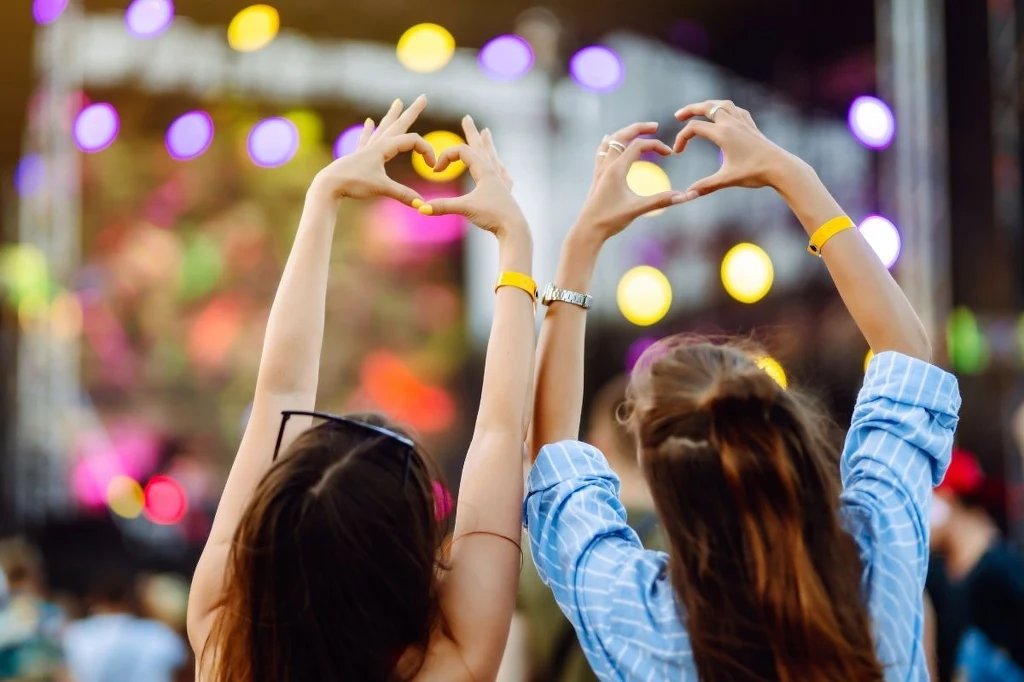 Koncert? Świetny pomysł! Dwie młode kobiety stojące w tłumie na koncercie plenerowym trzymają ręce uniesione do góry i układają dłonie w kształt serca, kolorowe światła rozświetlają scenę w tle.