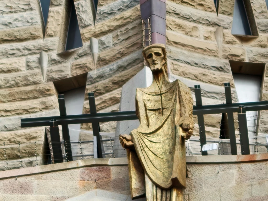 Sagrada Familia Złoty posąg postaci o surowym wyrazie twarzy, ustawiony na tle kamiennej fasady z geometrycznymi oknami. W tle charakterystyczna architektura z ostrymi kątami i oryginalnym wzornictwem.