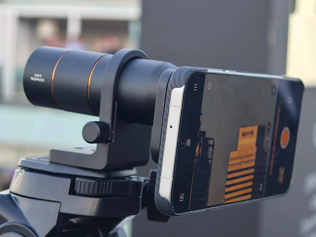 Telekonwerter do smartfona Oppo Find X9 Pro. Smartfon zamocowany na statywie z zewnętrznym obiektywem teleskopowym umożliwiającym wykonywanie zdjęć w większym przybliżeniu. W tle widoczne rozmyte elementy otoczenia.