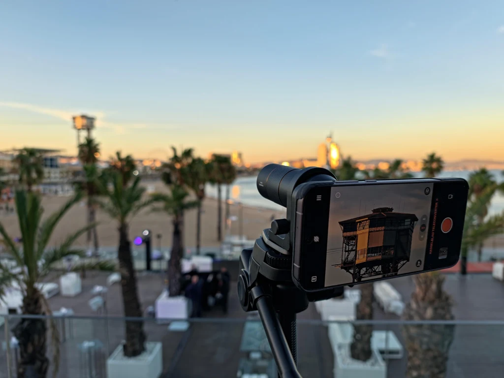 Zbliżenie telekonwerterem na wieżę kolejki w Barcelonie. Miejsca dzieliło około 700 metrów. Smartfon zamocowany na statywie wykonuje zdjęcie nadmorskiego krajobrazu z palmami i jasnym niebem, w tle widoczna piaszczysta plaża, nowoczesne budynki oraz rozstawione białe meble ogrodowe.