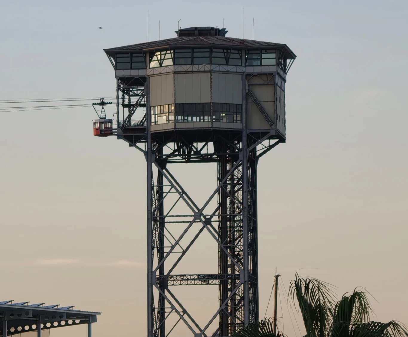 Wysoka metalowa wieża z przeszkloną górną częścią otoczona elementami konstrukcyjnymi, obok linia kolejki linowej z kabiną, u dołu fragment liści palmowych, błękitne niebo w tle.