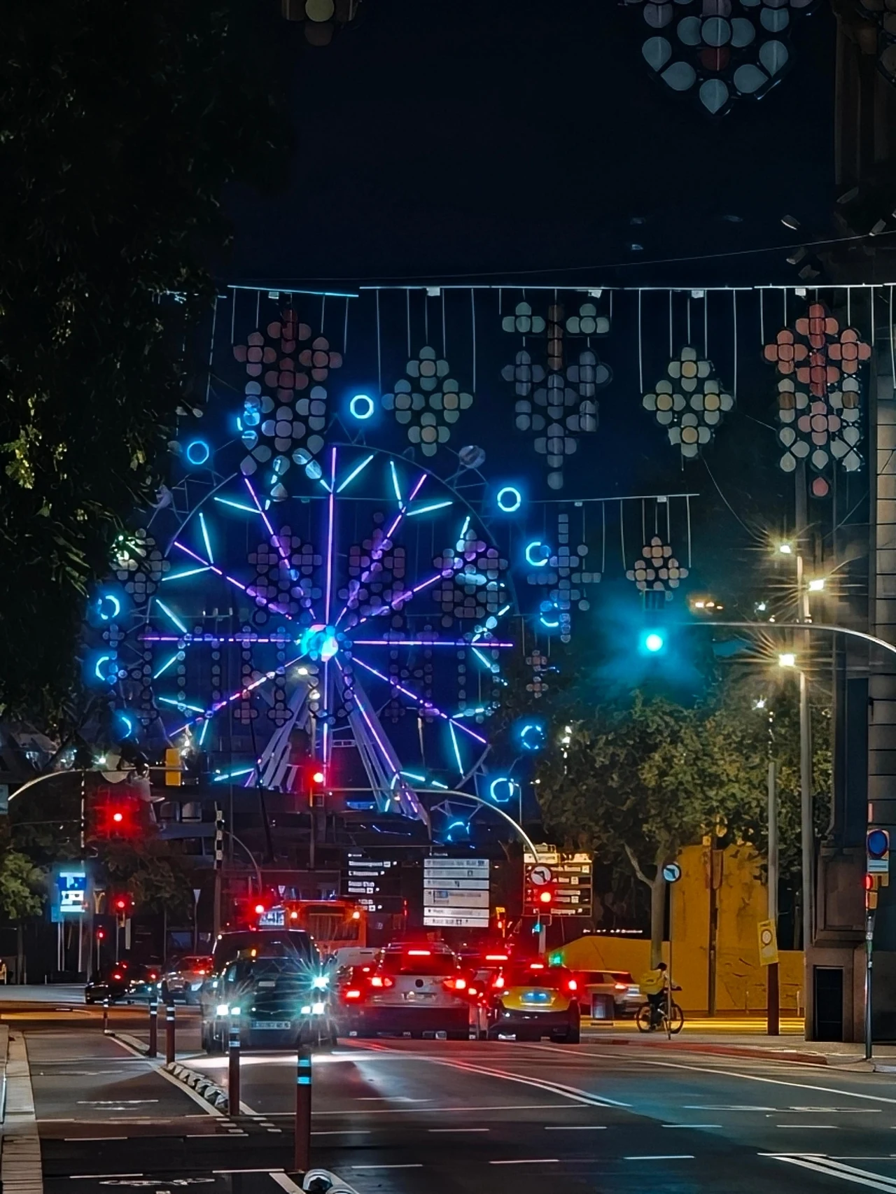 Barcelona na wieczornych i nocnych zdjęciach. Kolorowy diabelski młyn oświetlony neonami na tle miejskiej ulicy nocą, lampki zawieszone nad ulicą, liczne samochody oczekujące na światłach, widoczne drzewa po bokach jezdni.