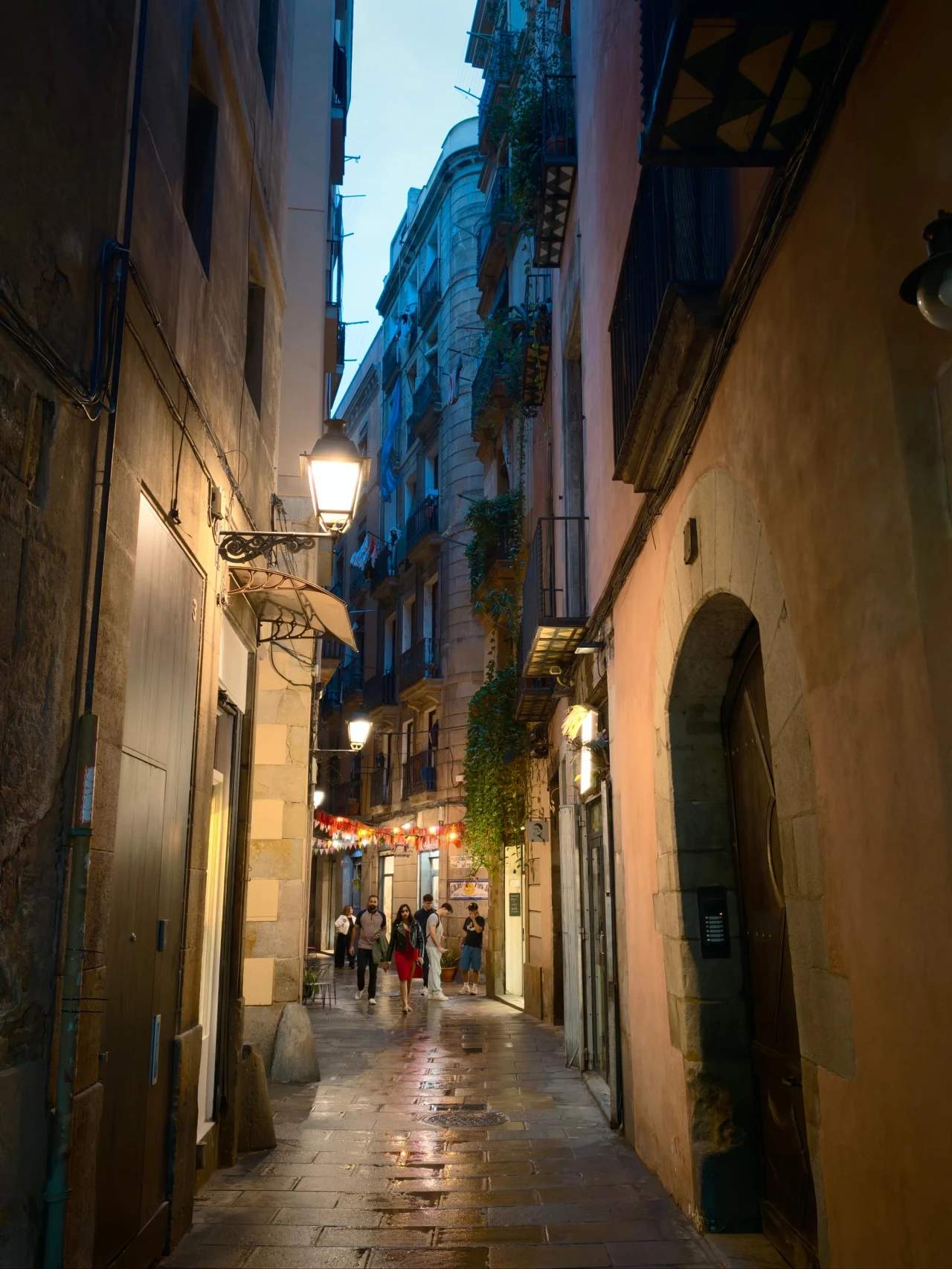 Barcelona na wieczornych i nocnych zdjęciach. Wąska uliczka w zabytkowej części miasta oświetlona latarniami, z wysokimi budynkami po obu stronach, licznymi balkonami i pnącą roślinnością. Na końcu ulicy widoczna grupa przechodniów oraz delikatne światła restauracji lub kawiarni, tworzące przytuln...