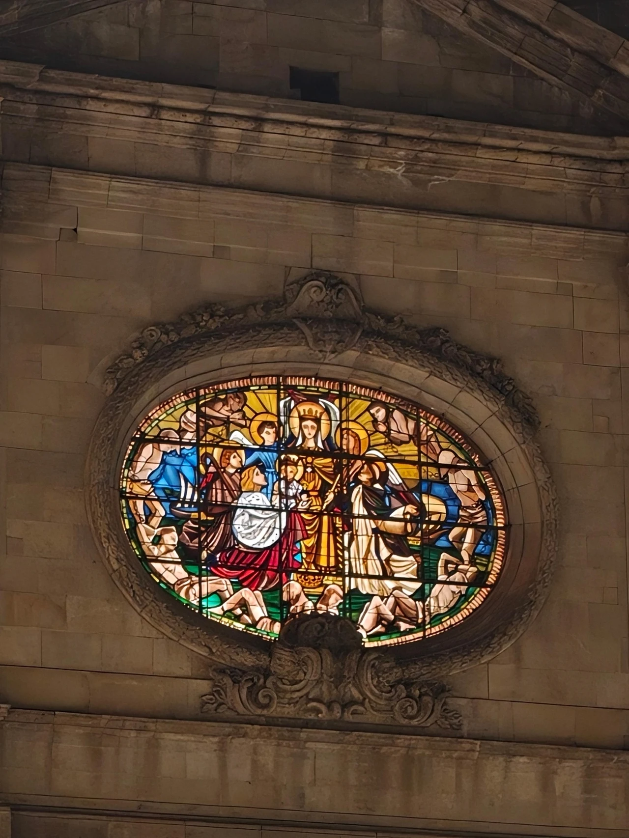 Barcelona na wieczornych i nocnych zdjęciach. Witraż w owalnym oknie przedstawiający postaci ubrane w szaty w scenerii religijnej, centralnie znajduje się kobieta z dzieckiem na ręku otoczona przez innych ludzi, całość oświetlona ciepłym światłem.