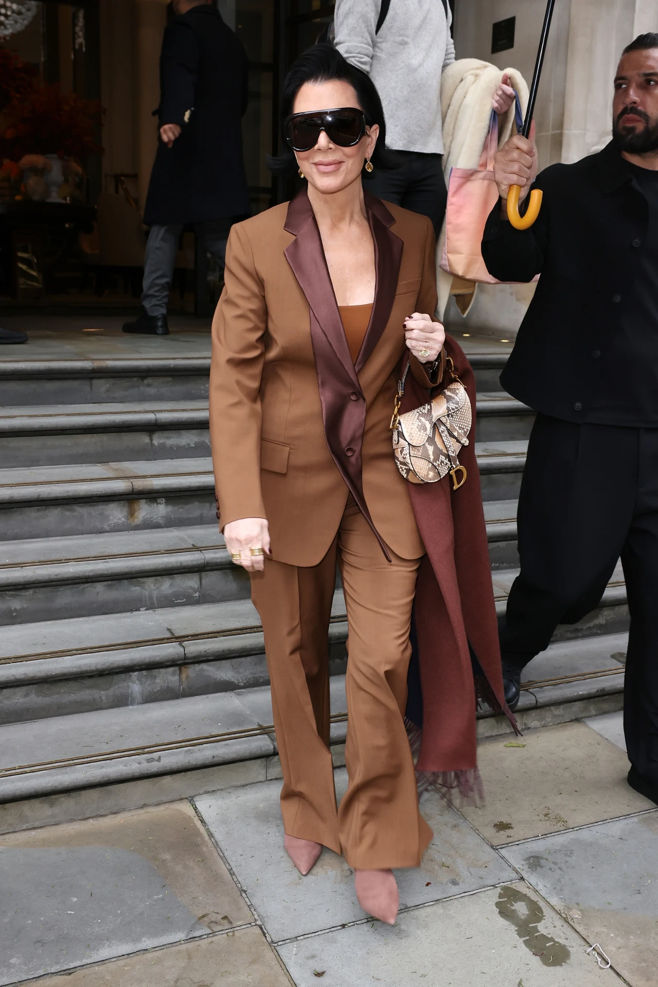 Kobieta ubrana w elegancki brązowy garnitur i okulary przeciwsłoneczne stoi na schodach przed budynkiem, trzymając modną torebkę ze wzorem węża. Za nią znajduje się kilku ludzi, jeden z nich trzyma parasol.