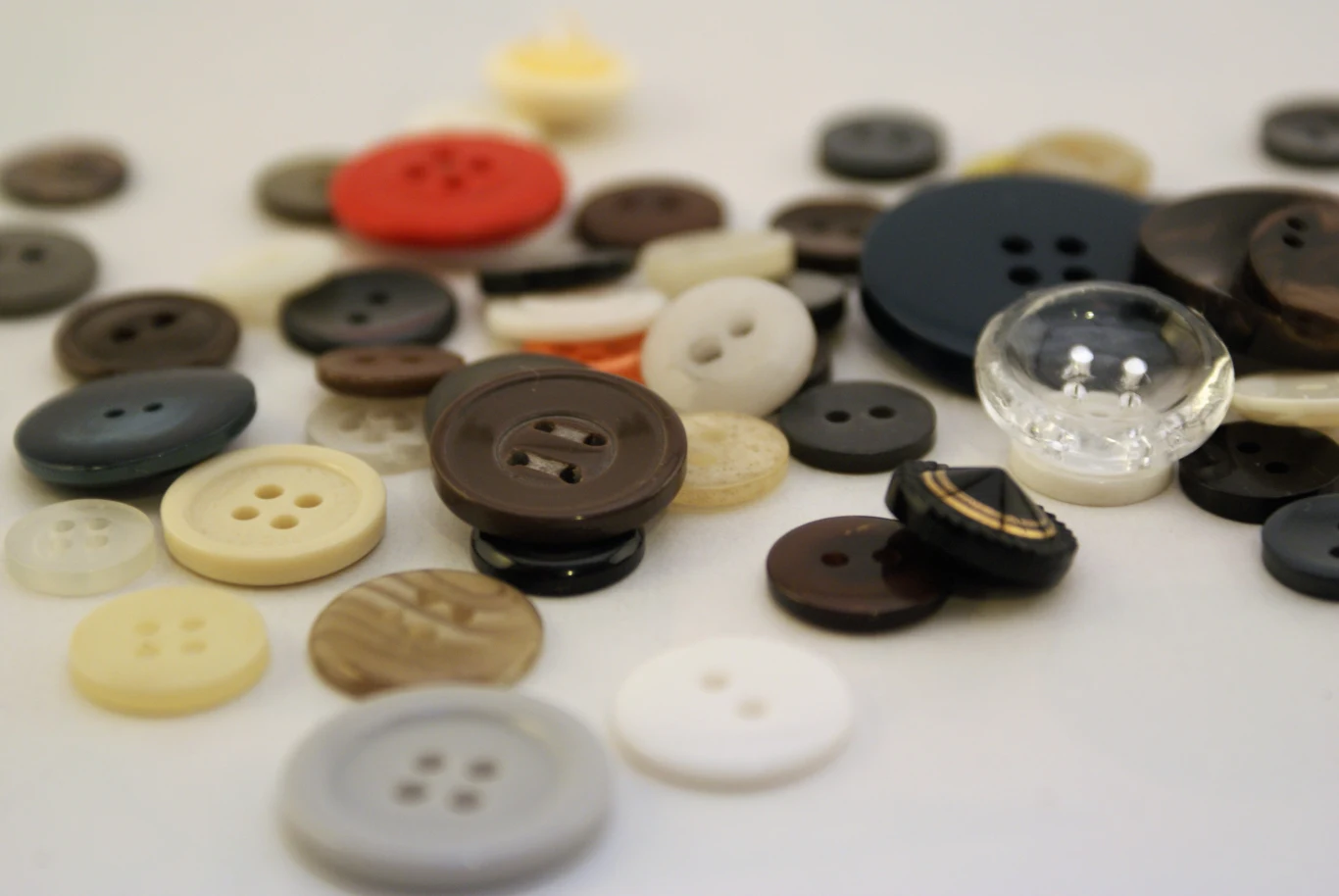 Różnorodne guziki w wielu kolorach, rozmiarach i kształtach rozrzucone na jasnej powierzchni, w tym guziki plastikowe, przezroczyste i drewnopodobne.