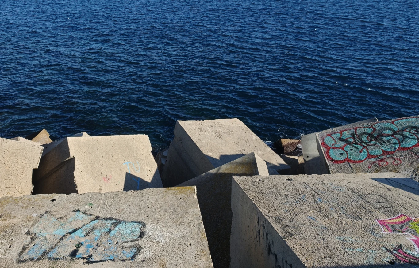 Betonowe bloki wyłożone przy brzegu morza, pokryte miejscami graffiti i napisami, z falującą wodą w tle.