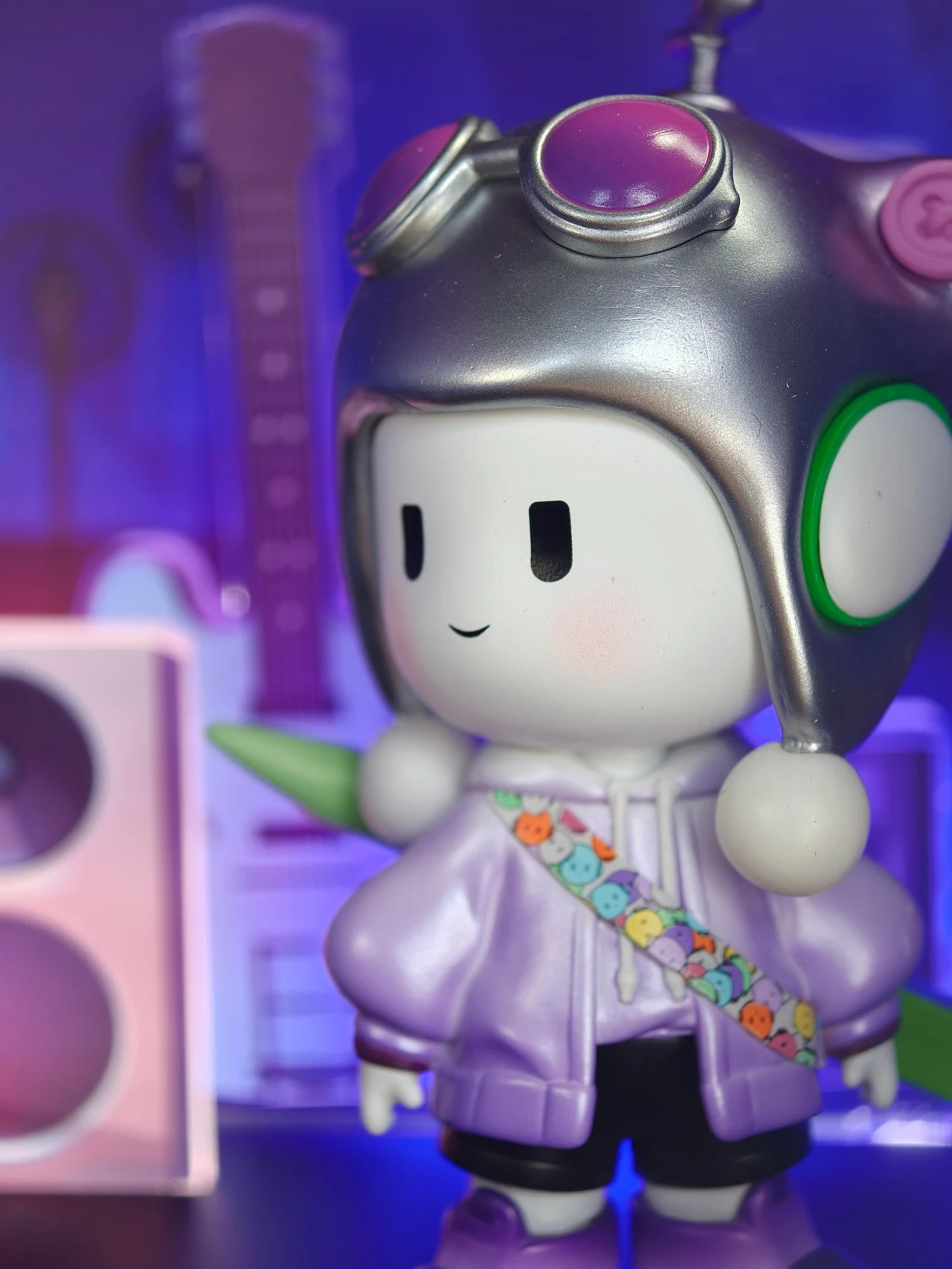 Zdjęcia zrobione przez Oppo Find X9 Pro. Kolorowa figurka o dużej głowie i prostych rysach twarzy ubrana w fioletową kurtkę z ozdobnym paskiem przedstawiającym buźki. Na głowie ma srebrny kask z różowymi i zielonymi elementami. W tle rozmyte, pastelowe kształty i elementy przypominające głośn...