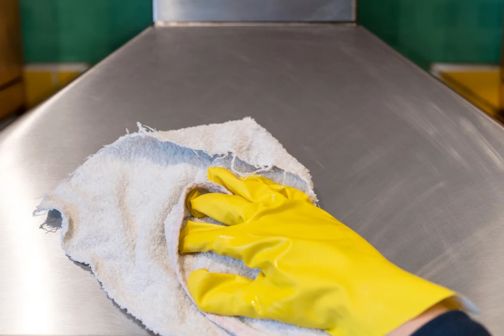 Idealnie czysty okap? Trzeba przecierać go codziennie Ręka w żółtej gumowej rękawiczce wyciera metalową powierzchnię białą, nieco postrzępioną szmatką.