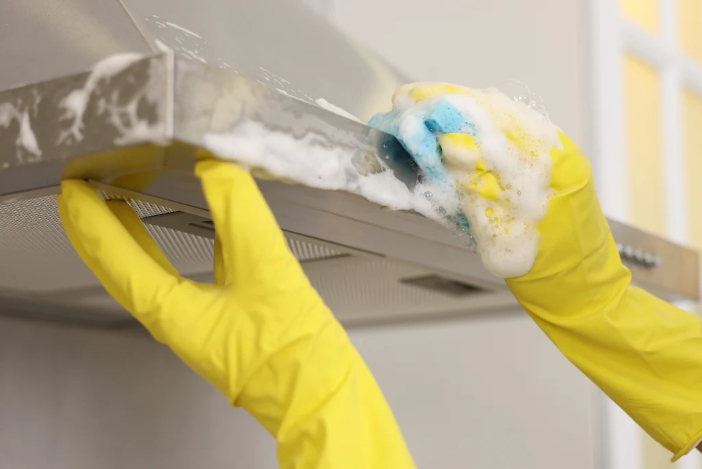 Płyn do mycia naczyń nie zawsze poradzi sobie z tłuszczem na okapie` Osoba w żółtych rękawiczkach czyści okap kuchenny za pomocą gąbki z pianą, skupiając się na usuwaniu zabrudzeń z powierzchni urządzenia.