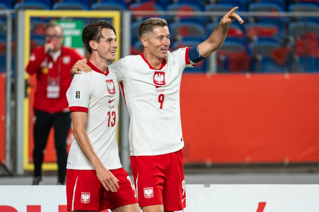 Jakub Kamiński (z lewej) Dwóch piłkarzy reprezentacji Polski w biało-czerwonych strojach cieszy się po meczu na stadionie, jeden obejmuje drugiego, a drugi wskazuje ręką coś w oddali; w tle rozmazane sylwetki i trybuny.