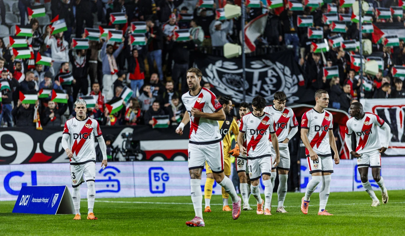 Piłkarze Rayo Vallecano w sezonie 2025/2026 Grupa piłkarzy ubranych w białe stroje z czerwoną szarfą biegnie po murawie stadionu, w tle na trybunach kibice z flagami pokazują wsparcie swojej drużynie.