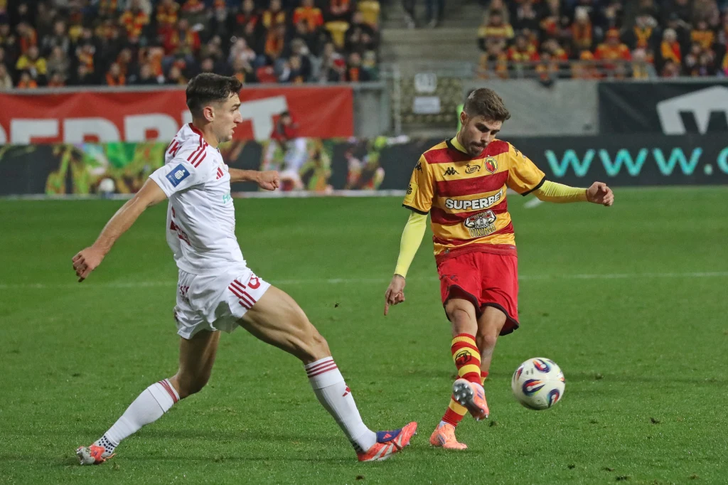 Jagiellonię czeka wyprawa przez całą Polskę, Raków Częstochowa uda się do Wrocławia Dwóch piłkarzy walczy o piłkę w trakcie meczu piłkarskiego, jeden w białym stroju próbuje zablokować zagranie rywala w żółto-czerwonym stroju, w tle kibice na trybunach.