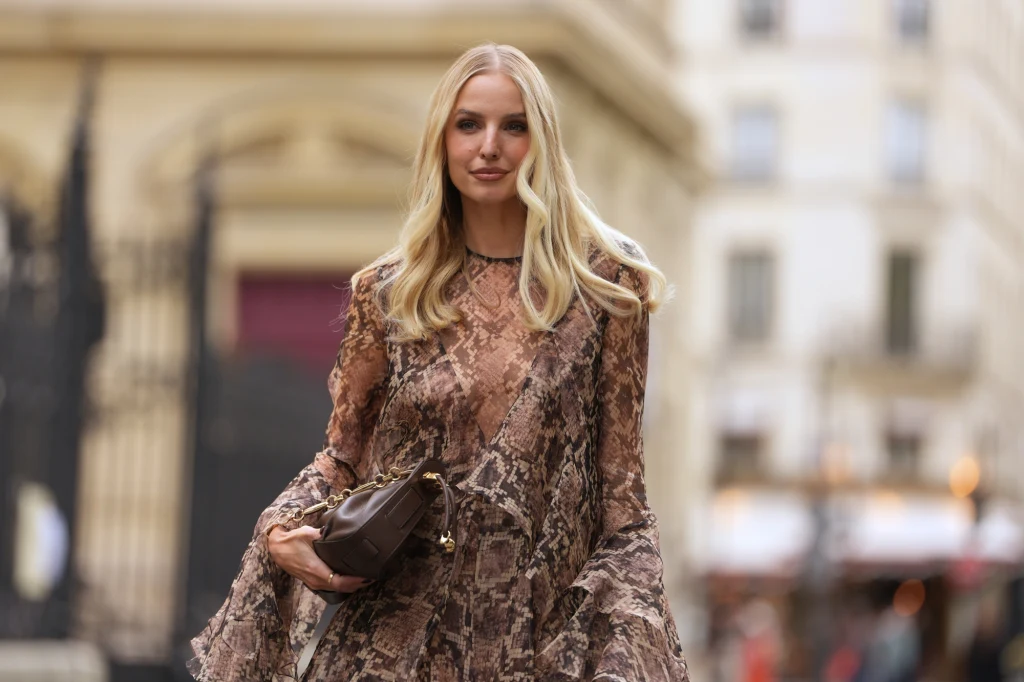 Jak dobierać kolory, aby podkreślić swój typ urody? Kobieta o blond włosach ubrana w długą sukienkę o motywie wężowej skóry stoi na tle miejskiej architektury i trzyma w rękach brązową kopertówkę.