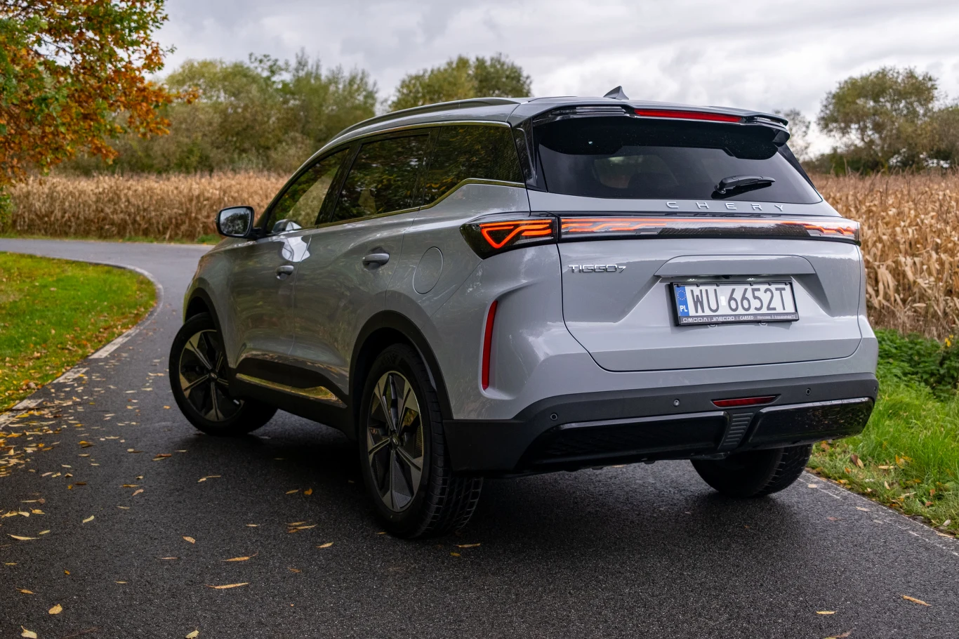 Chery Tiggo 7 to konstrukcyjny bliźniak Jaecoo 7 - wywodzi się zresztą z tego samego koncernu i ma ten sam układ napędowy. Srebrny SUV zaparkowany na asfaltowej drodze wśród jesiennych drzew i pól kukurydzy, widok od tyłu pod kątem, wyraźnie widoczna tablica rejestracyjna i charakterystyczne tylne światła.