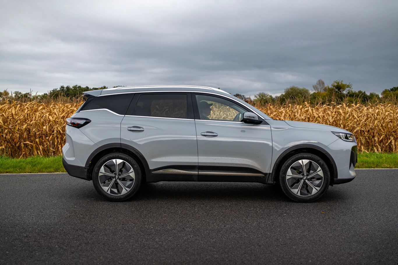 Na długość nadwozia Tiggo przerasta swoich rywali, ale już rozstawem osi nieznacznie im ustępuje. Srebrny samochód typu SUV stoi zaparkowany na asfaltowej drodze, w tle znajduje się pole kukurydzy, a niebo jest pochmurne.
