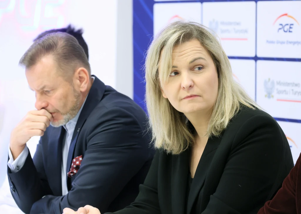 Otylia Jędrzejczak Dwie osoby siedzą przy stole konferencyjnym, kobieta w jasnych włosach w ciemnej marynarce patrzy w bok, mężczyzna w garniturze opiera dłoń o twarz. W tle widoczne plansze z logotypami i napisami.