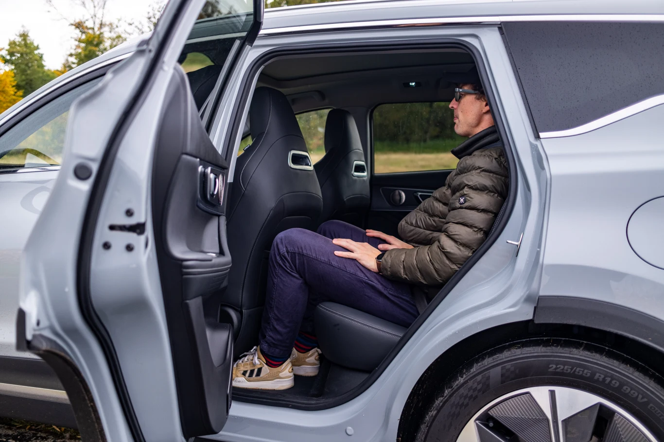 Przestrzeni na kanapie nie brakuje. Fotele ze zintegrowanymi zagłówkami z przodu to czysta estetyka - ich trzymanie boczne jest słabiutkie. Mężczyzna siedzi na tylnym siedzeniu nowoczesnego samochodu, drzwi pojazdu są otwarte, a wnętrze auta prezentuje się przestronnie. Ubrany jest w ciemne spodnie, sportową kurtkę i adidasy, patrzy przed siebie.