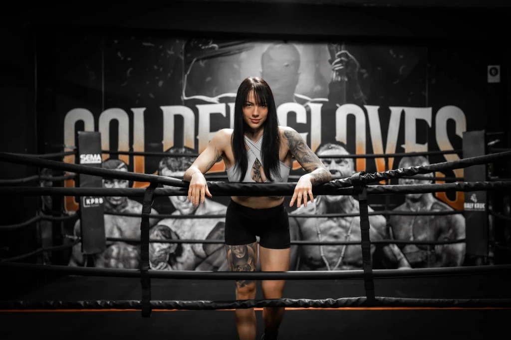 Adrianna Jędrzejczyk Kobieta w sportowym stroju stoi w ringu bokserskim, opierając się o liny, w tle widoczne są plakaty z wizerunkami bokserów i napis 'Golden Gloves', całość utrzymana w siłownianej atmosferze.