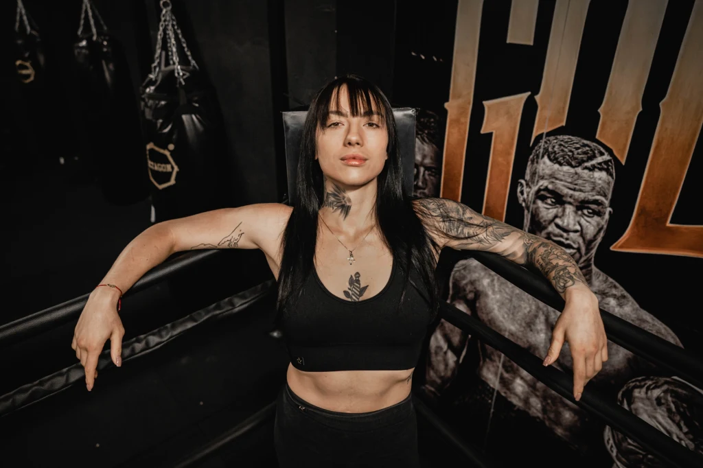 Adrianna Jędrzejczyk Silna kobieta w sportowym stroju stojąca w ringu bokserskim z odsłoniętymi, umięśnionymi ramionami, widoczne tatuaże na ciele oraz mural boksera na ścianie w tle.