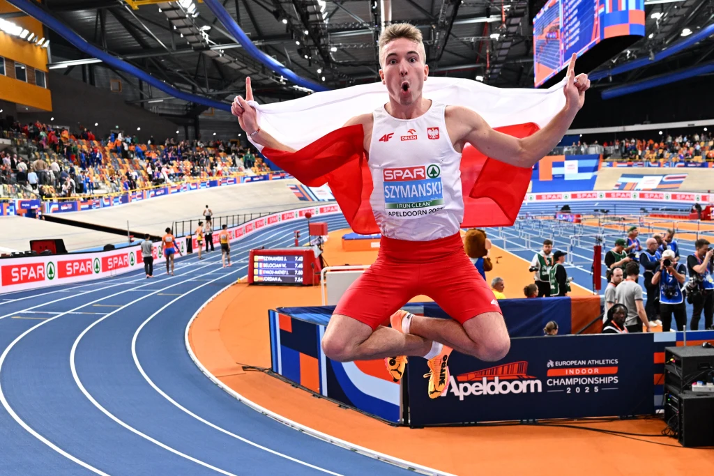 Jakub Szymański po sukcesie w HME w Apeldoorn Sportowiec w czerwono-białym stroju unosi się w powietrzu po skoku, trzymając szeroko rozpostartą polską flagę. W tle widać bieżnię lekkoatletyczną, kibiców oraz oznaczenia zawodów halowych. Panuje atmosfera radości i sukcesu.