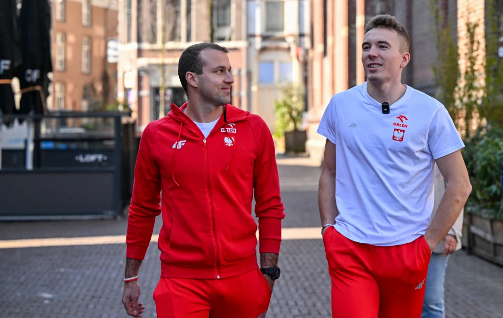 Jakub Szymański (po prawej) z trenerem Mikołajem Justyńskim Dwóch mężczyzn ubranych w sportowe stroje z polskimi emblematami idzie chodnikiem miejskim, uśmiechając się i rozmawiając ze sobą; w tle zabudowa miejska i światło słoneczne.