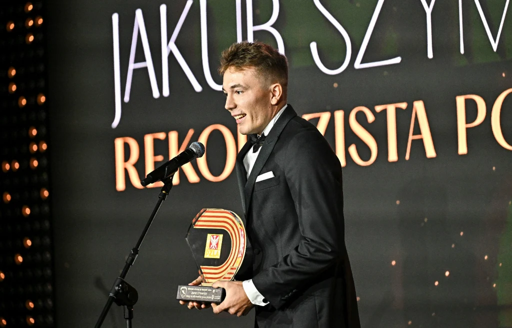 Jakub Szymański na gali "Złote Kolce" Mężczyzna w smokingu odbierający nagrodę na scenie, trzymając statuetkę i przemawiając do mikrofonu. W tle widoczny jest podświetlony napis z imieniem i nazwiskiem oraz tytułem wyróżnienia.