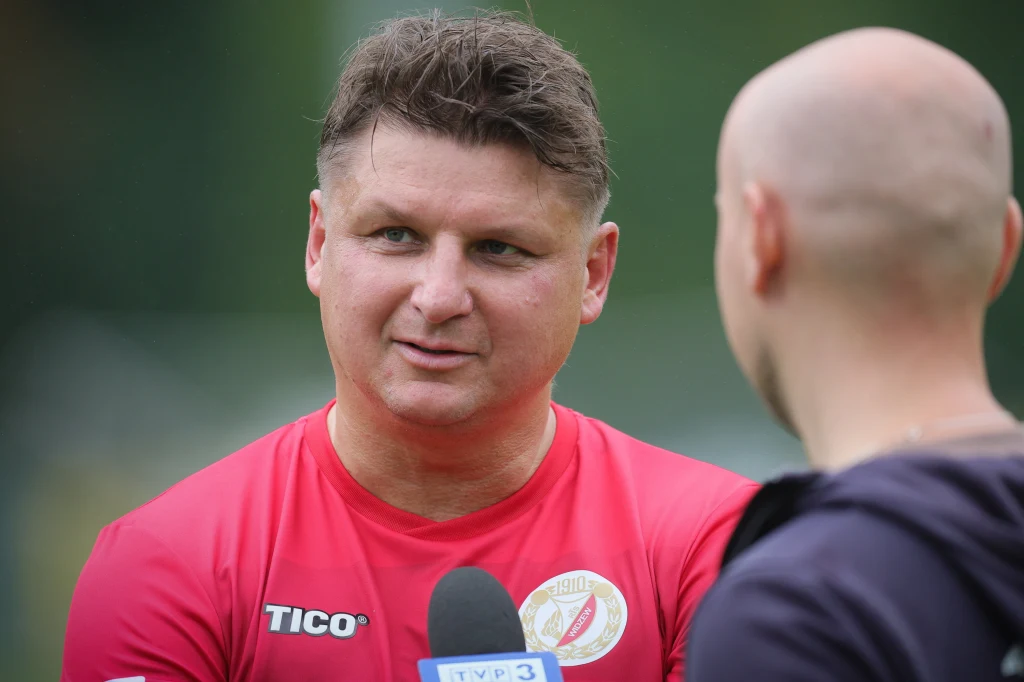 Mężczyzna w czerwonej koszulce sportowej udziela wywiadu przed mikrofonem z logo TVP3, w tle rozmyty zielony krajobraz.