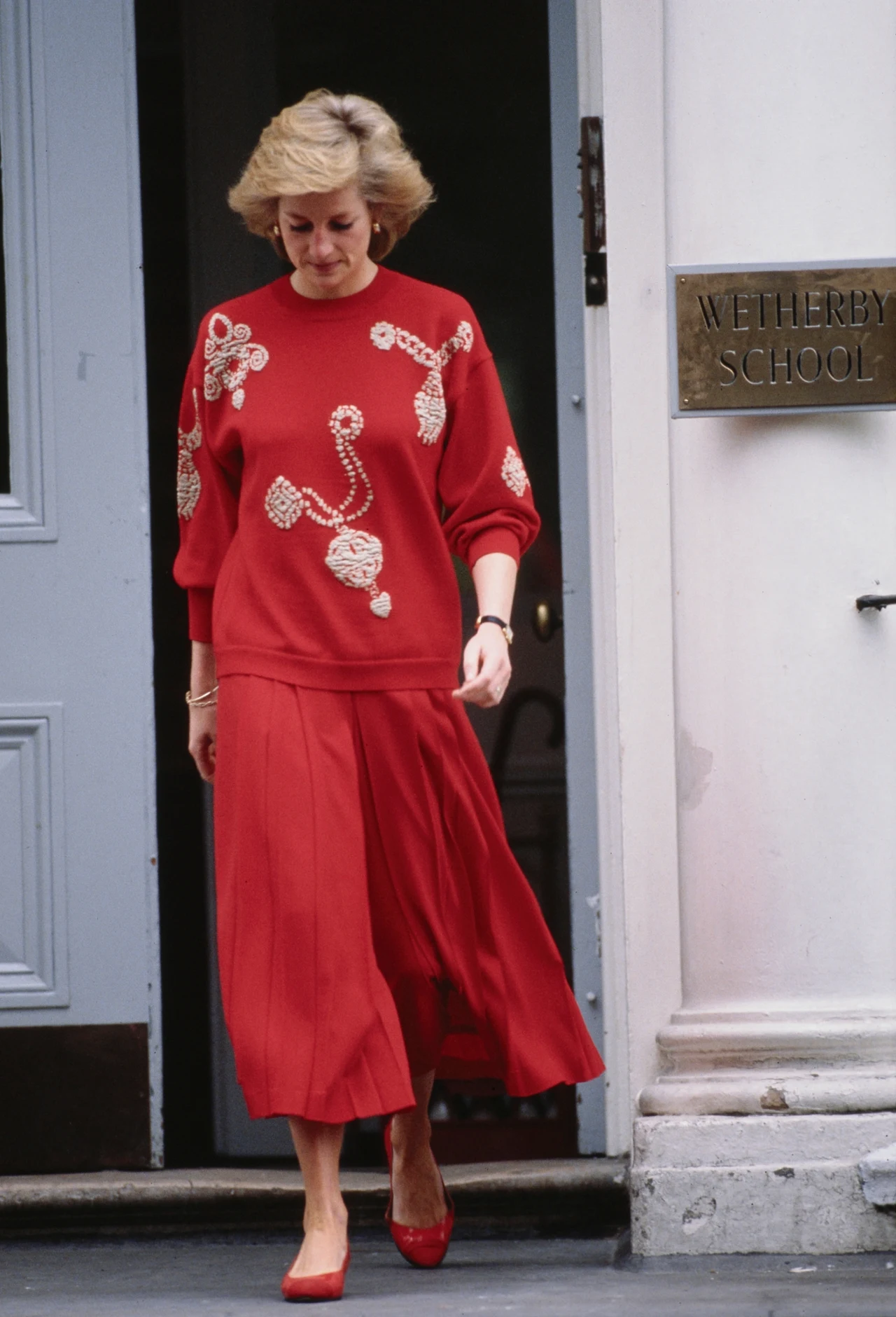 Kobieta ubrana w czerwony sweter z ozdobnymi aplikacjami oraz czerwoną, plisowaną spódnicę wychodzi z budynku oznaczonego tabliczką Wetherby School.