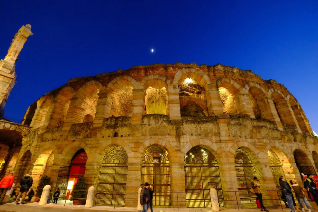 Średniowieczny amfiteatr w Weronie będzie areną ceremonii zamknięcia zimowych igrzysk olimpijskich 2026 Starożytny amfiteatr oświetlony żółtym światłem na tle głębokiego, niebieskiego nieba. Kręgi łuków i kolumn tworzą monumentalną budowlę w centrum, wokół której przechodzą ludzie.