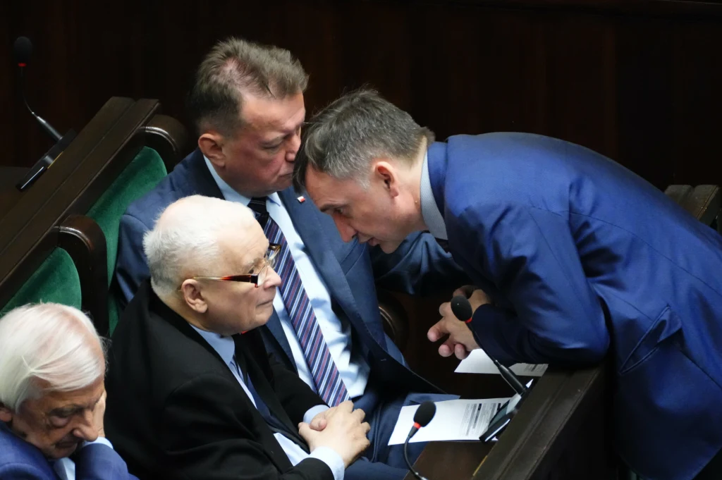 Jarosław Kaczyński i Zbigniew Ziobro w Sejmie Grupa mężczyzn w garniturach podczas rozmowy w ławach parlamentu, jeden z nich nachyla się i prowadzi konwersację z pozostałymi.