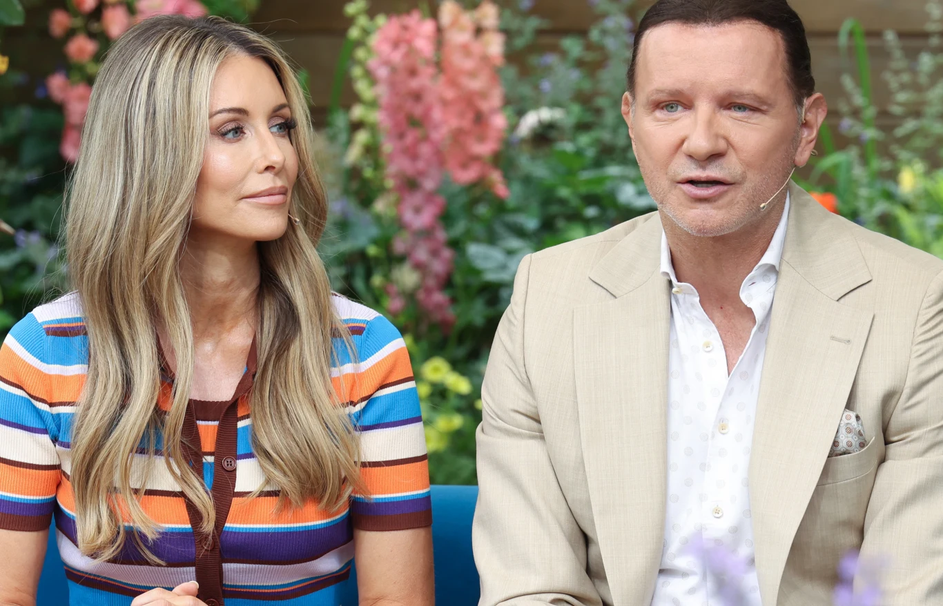 Dwoje dorosłych siedzi obok siebie na tle kolorowych kwiatów i zielonych roślin. Kobieta ma długie blond włosy i kolorowy pasiasty sweter, mężczyzna zaś jasny garnitur i białą koszulę. Oboje wyglądają na zaangażowanych w rozmowę, mają zamocowane mikrof...