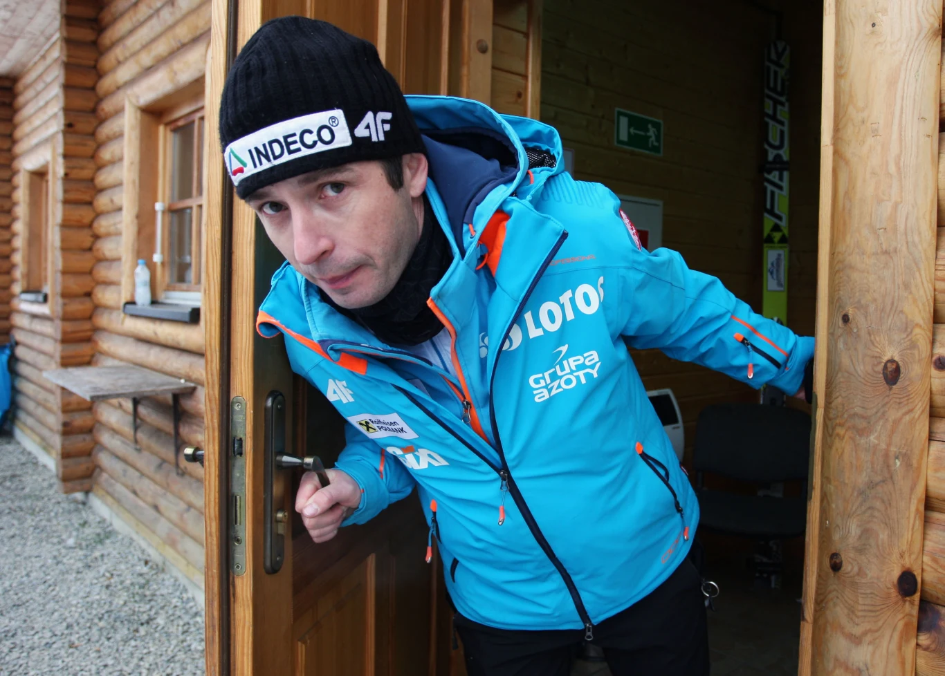 Łukasz Kruczek w 2014 roku Mężczyzna w niebieskiej kurtce sportowej z logotypami sponsorów wychodzi z drewnianego budynku, nosi czapkę z logo i spogląda w stronę aparatu, otoczenie sugeruje górski lub zimowy charakter wydarzenia.
