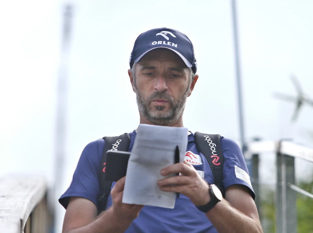 Łukasz Kruczek Mężczyzna w sportowej koszulce i czapce z logo marki Orlen czyta dokumenty, trzymając otwarte etui, w tle widoczne rozmyte elementy architektury.