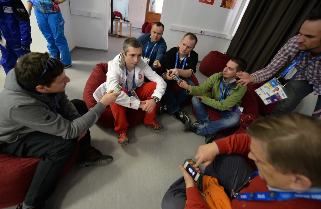 Łukasz Kruczek z dziennikarzami w czasie zimowych igrzysk olimpijskich w Soczi (2014) Grupa mężczyzn siedzących w kółku na pufach, kilku z nich trzyma dyktafony i telefony skierowane w stronę młodego mężczyzny w biało-czerwonym stroju sportowym, który jest w centrum uwagi, wygląda na udzielającego wywiad.